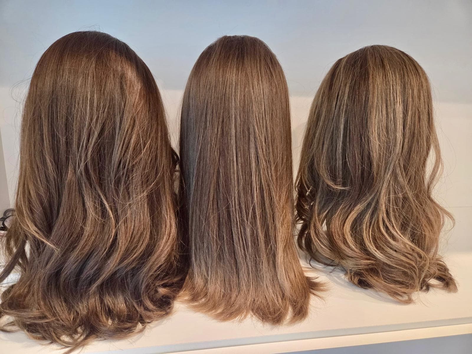 Three brown wigs in straight and wavy styles displayed from behind on a shelf.