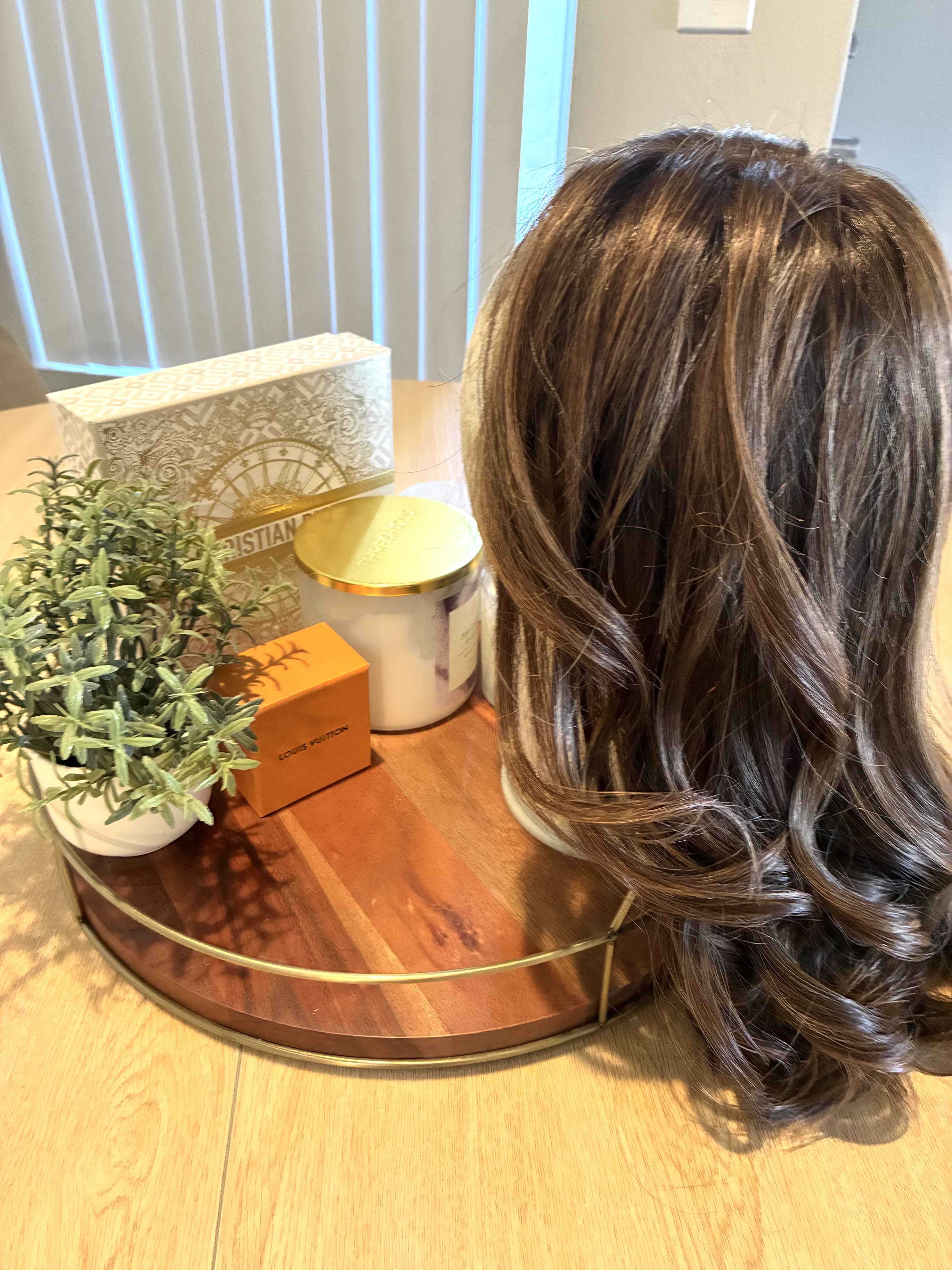 Brown wavy wig on mannequin head next to luxury brand boxes on a wooden tray.