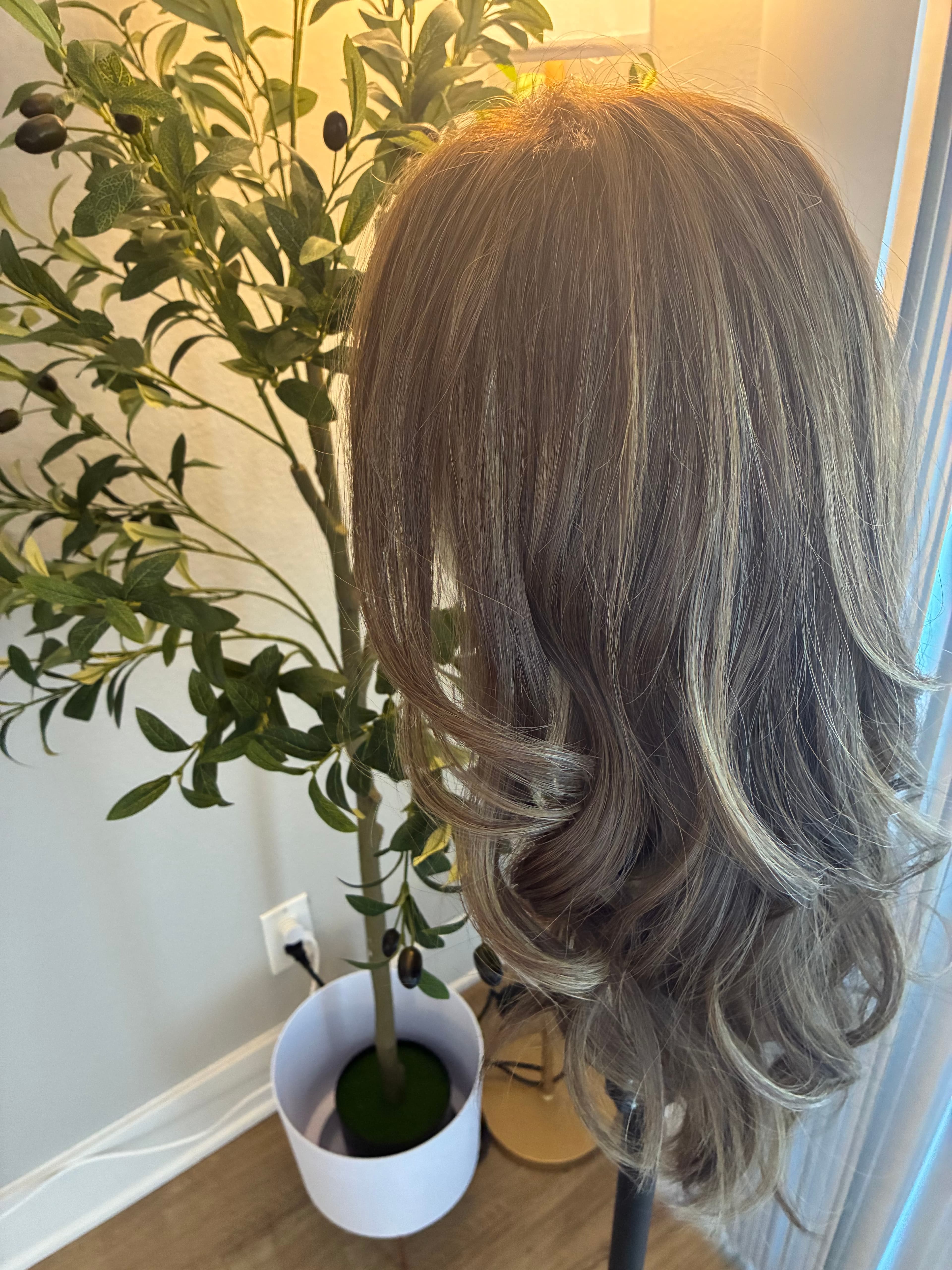 Long wavy ash-blonde wig on a stand next to a potted faux olive tree.