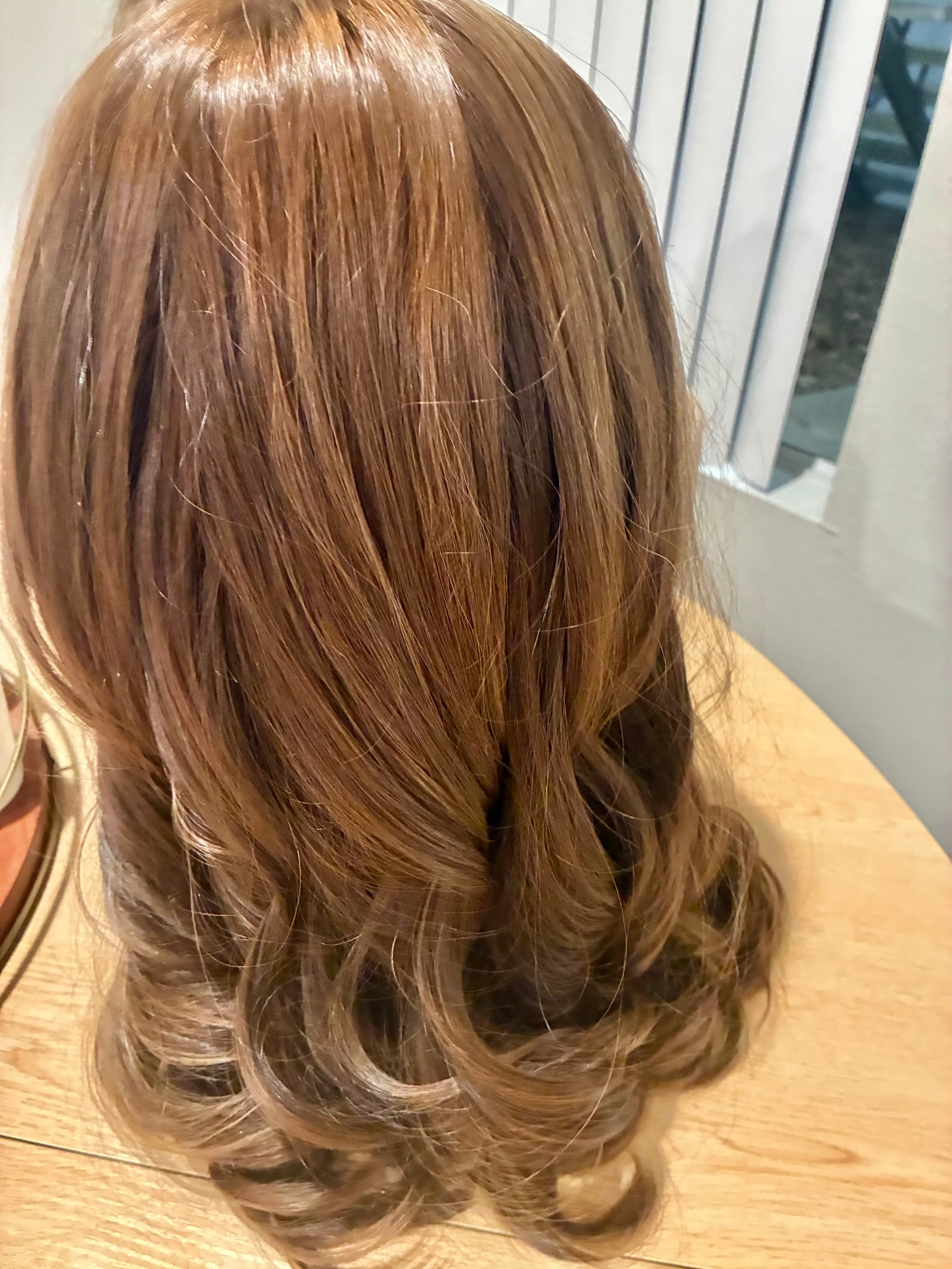 Long light brown wig with loose curls at the ends, displayed on a wooden table.